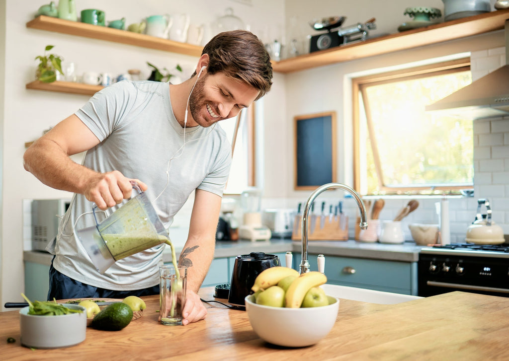 Man in kitchen wearing headphones, smiling while making a green smoothie